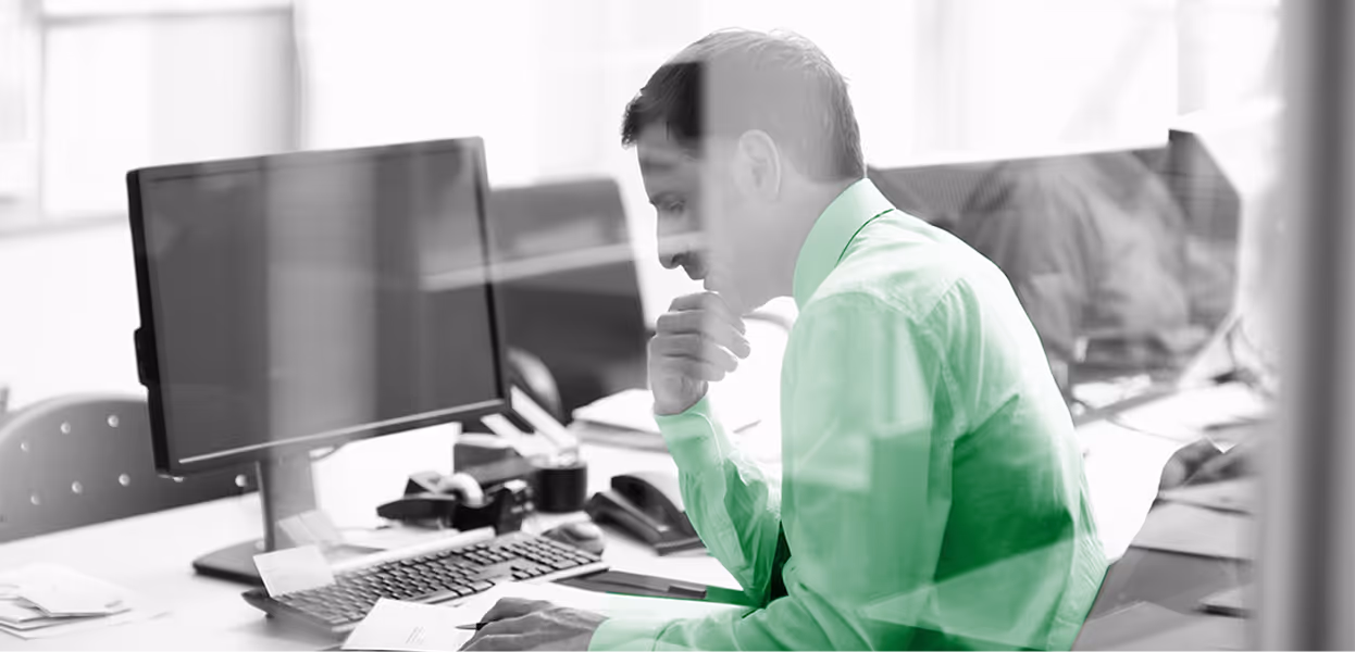 Man in a green shirt sitting at a desk, focused on his computer in a modern office setting.