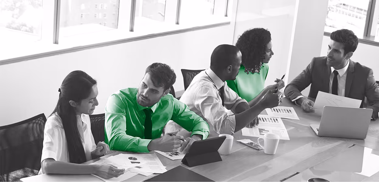 Group of five professionals in a meeting around a conference table discussing documents and using laptops, three wearing green highlighted clothing.