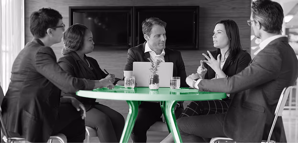 Five business professionals engaged in a meeting around a green round table, with a laptop and glasses of water present.