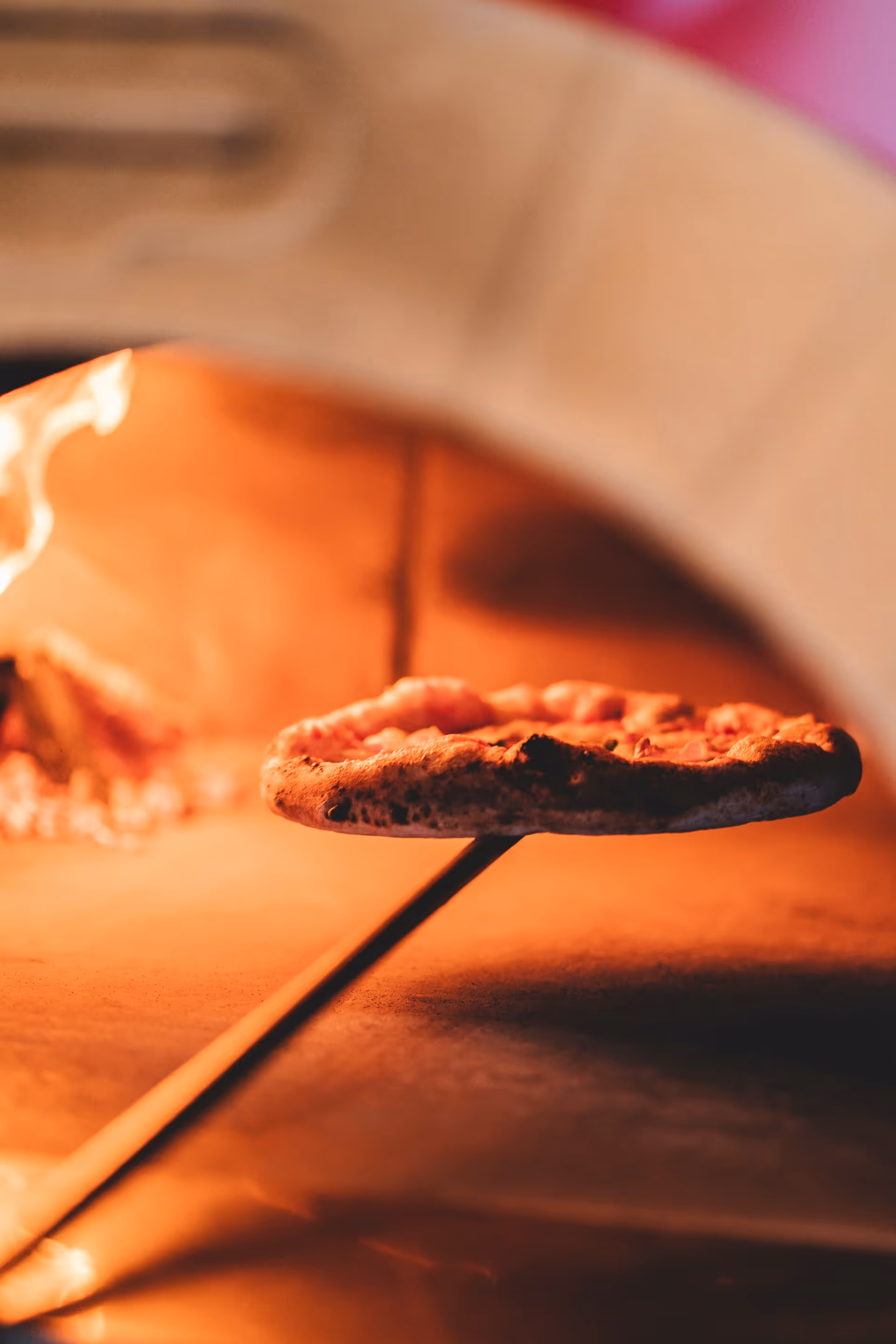 Pizza being cooked on a wooden peel in a rustic stone oven, with flames glowing warmly.