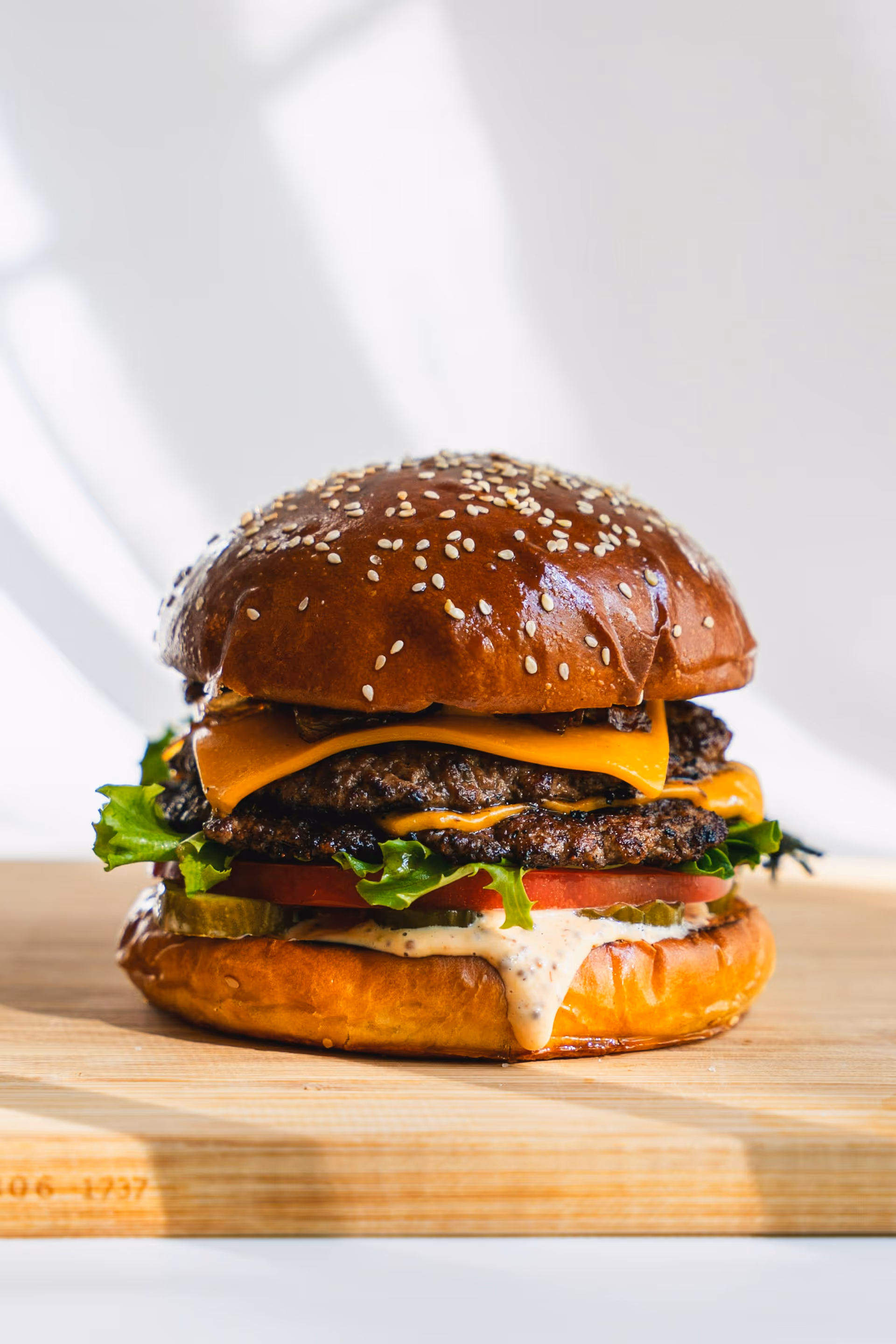 Juicy cheeseburger with a sesame bun, two beef patties, melted cheese, lettuce, tomato, pickles, and sauce, on a wooden board with soft lighting.