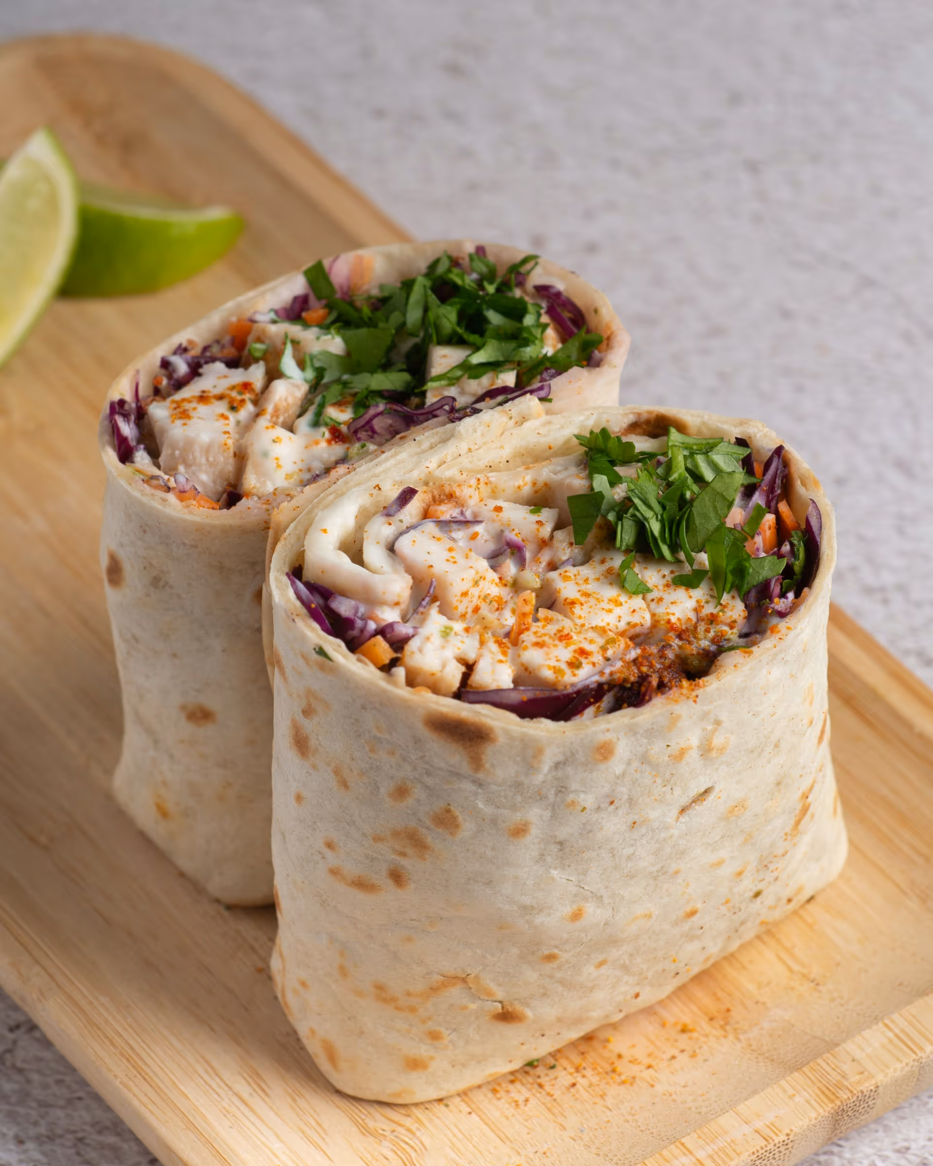 Two chicken wraps on a wooden board, filled with grilled chicken, purple cabbage, greens, and sprinkled with spices. Lime wedges are in the background.