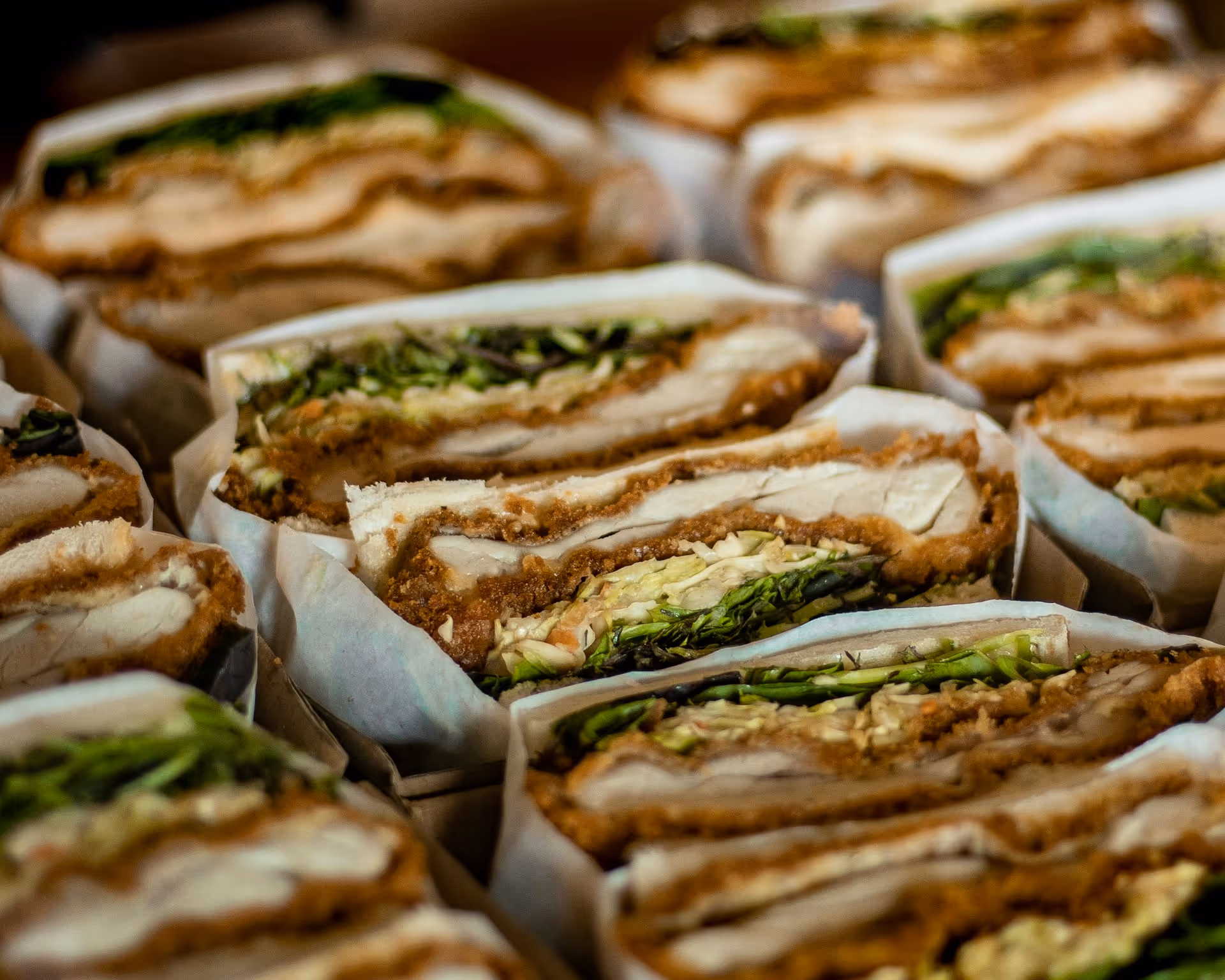 Close-up of multiple sandwiches filled with crispy chicken, fresh lettuce, and sauce, sliced in half and wrapped in paper.
