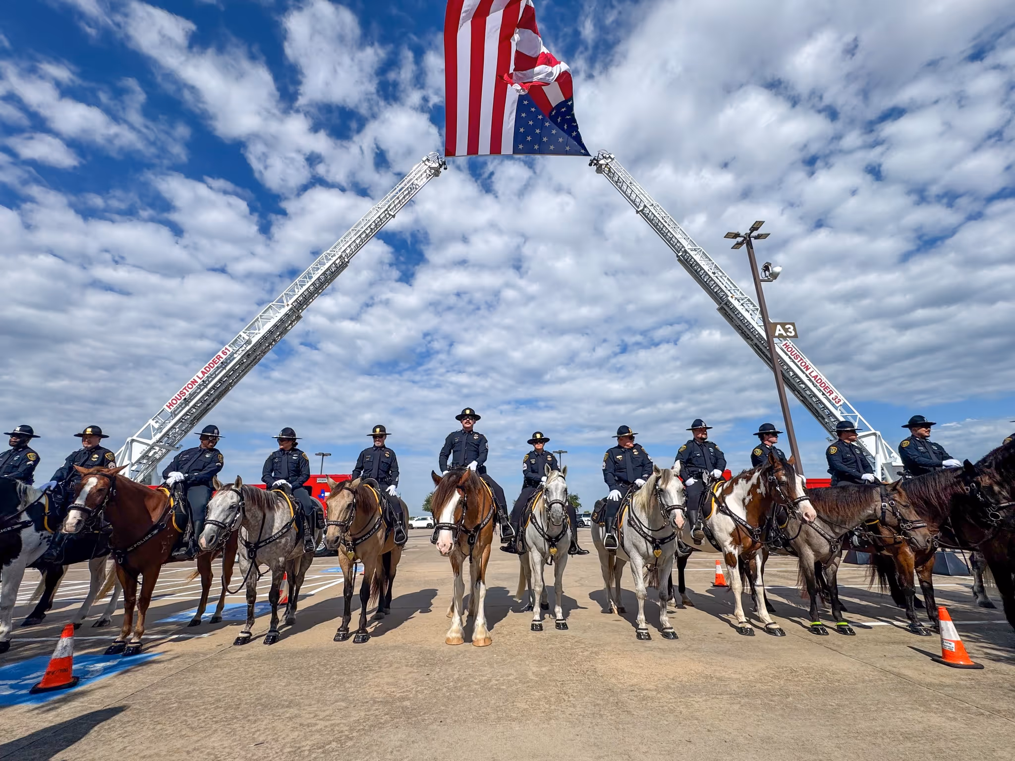 Mounted officers lined up on horseback beneath a large American flag suspended between two ladder trucks.