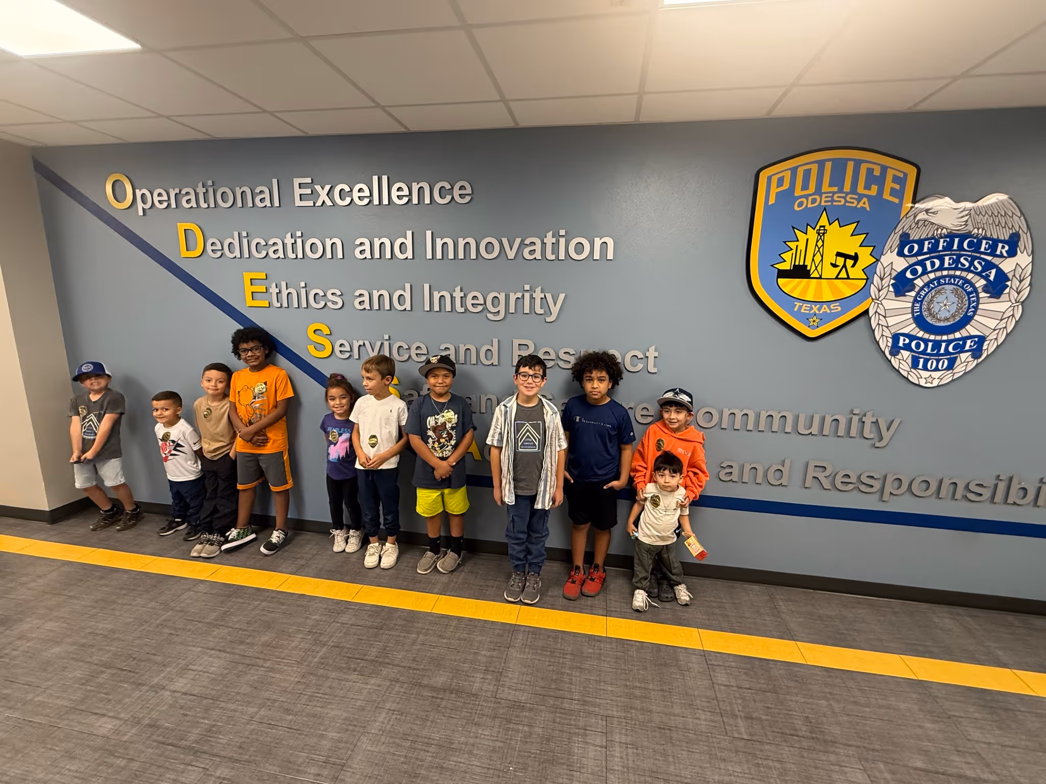 Group of children standing inside the Odessa Police Department in front of a wall displaying department values and badges.