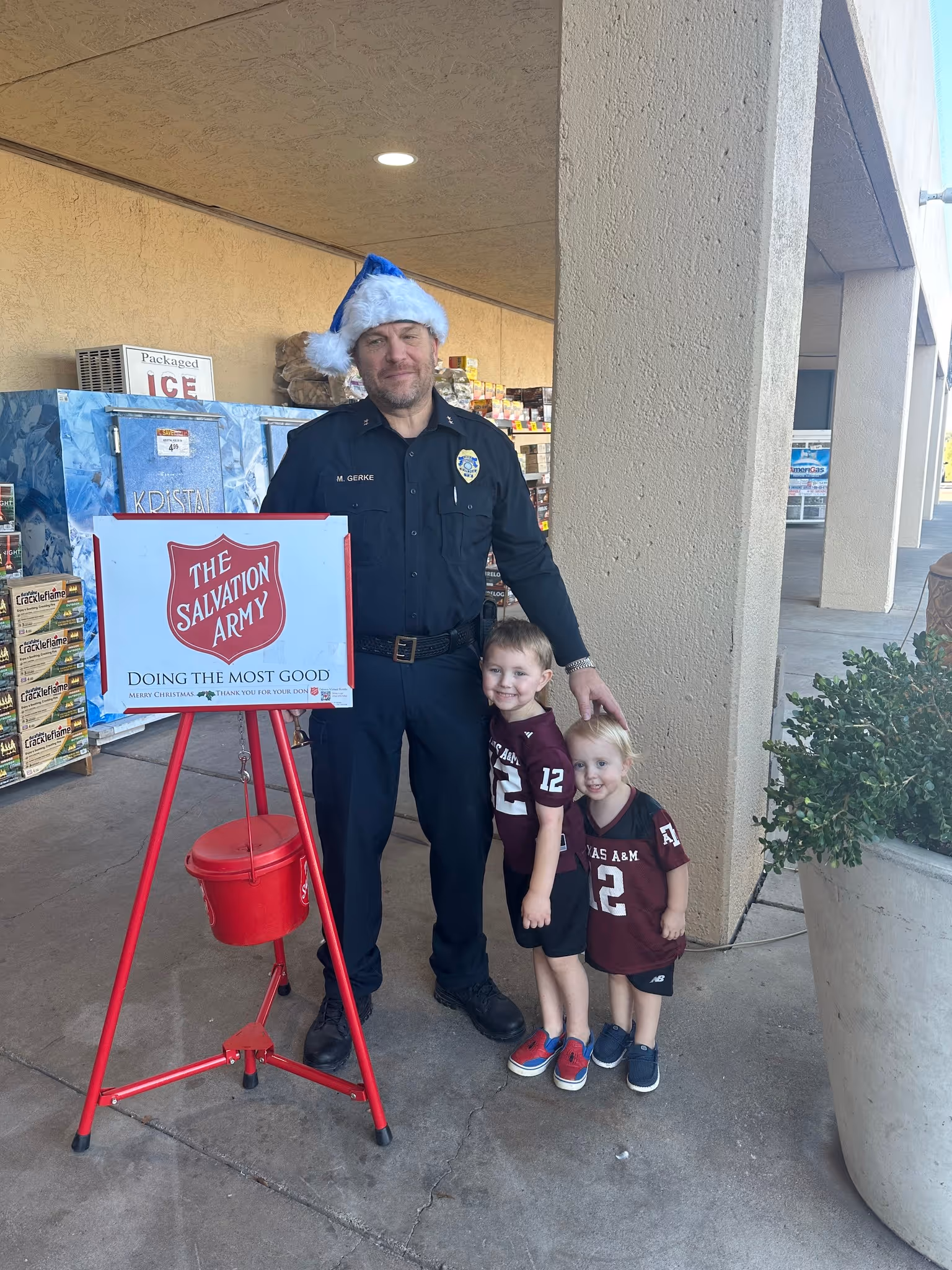 Officer in a Santa hat standing with two young children beside a Salvation Army donation kettle outside a store.