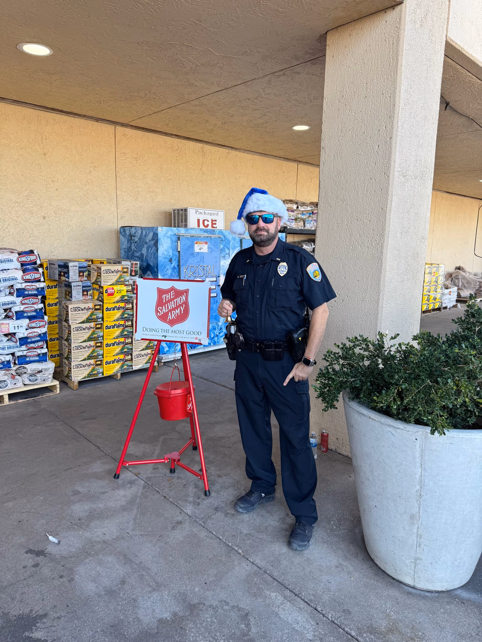 Officer wearing a Santa hat standing beside a Salvation Army donation kettle outside a storefront.