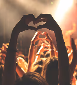 Silhouettes of people at a concert making heart shapes with their hands against bright stage lights.