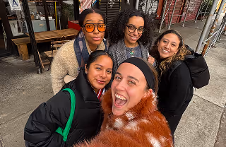 Five smiling women posing for a selfie outdoors near a construction site with scaffolding and wooden planks visible.
