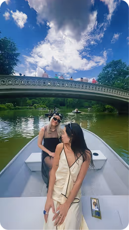 Two women sitting on a boat under a partly cloudy sky with a decorative bridge and trees in the background.