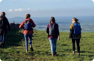 Four hikers with backpacks walking on a grassy hill with a clear sky and distant landscape.