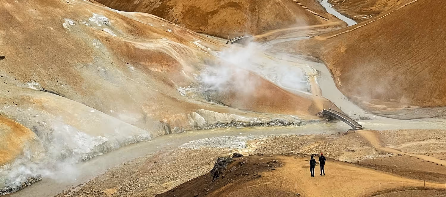 Two people walk on a trail in a rocky geothermal area with steam vents and a small river.