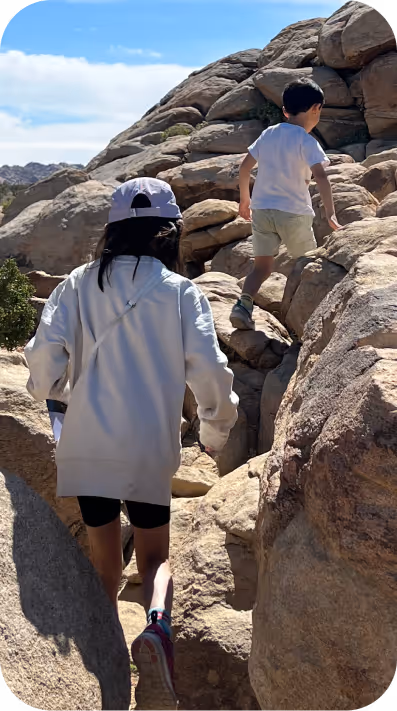 Two children climbing large rocks outdoors under a partly cloudy sky.