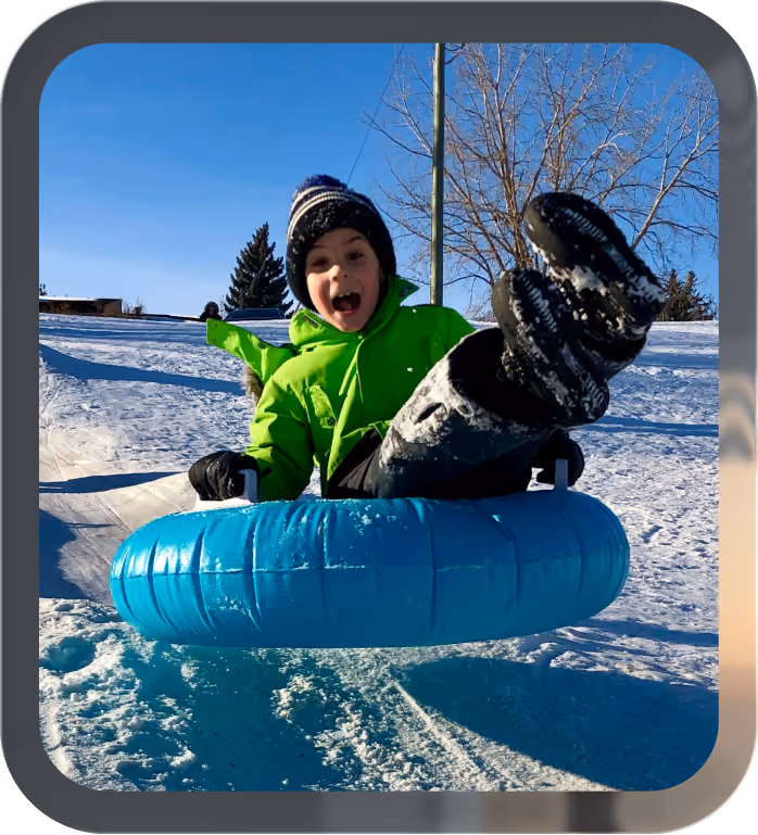 Child in bright green jacket and black hat snow tubing down a snowy hill on a blue tube.