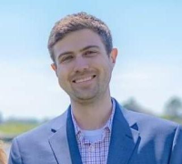 Smiling man with short brown hair wearing a blue blazer and checked shirt outdoors.