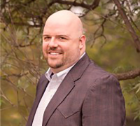 Smiling bald man in a dark blazer and white shirt standing outdoors with trees in the background.