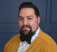 Man with dark hair and beard wearing a mustard yellow cardigan, white shirt with blue patterns, and a black bow tie, posing against a dark blue paneled wall.