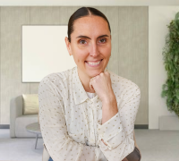 Smiling woman with dark hair tied back, resting her chin on her hand in a modern living room.