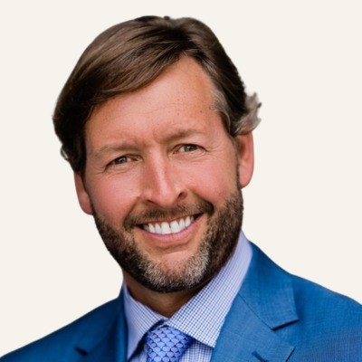 Smiling man with brown hair and beard wearing a blue suit and tie against a light background.