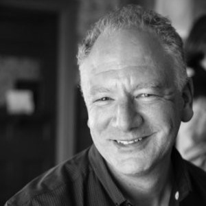 Black and white portrait of a smiling middle-aged man with curly gray hair wearing a dark shirt.