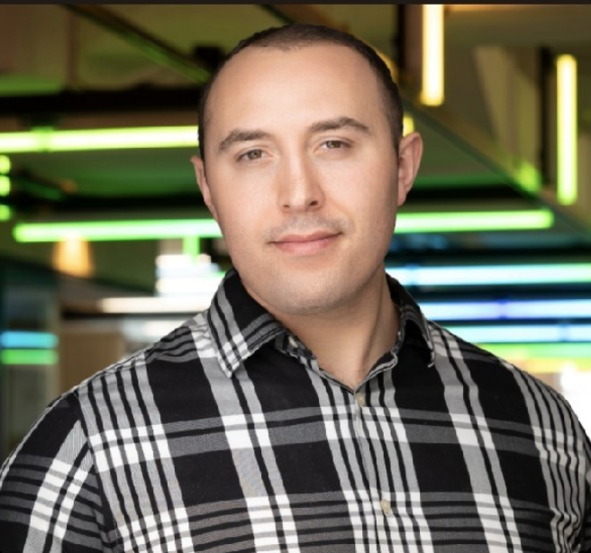 Portrait of a man with short dark hair wearing a black and white plaid shirt, standing indoors with blurred colorful lights in the background.