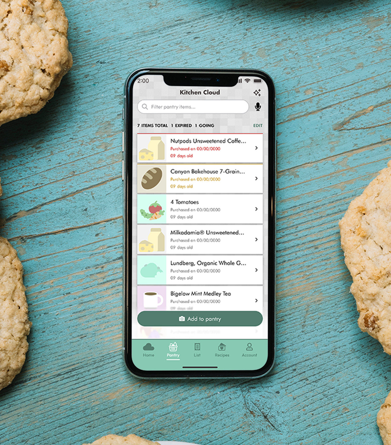Smartphone displaying a Kitchen Cloud app pantry list with items like Nutpods unsweetened coffee creamer, Canyon Bakehouse bread, tomatoes, and Bigelow mint tea, placed on a blue wooden surface surrounded by cookies.