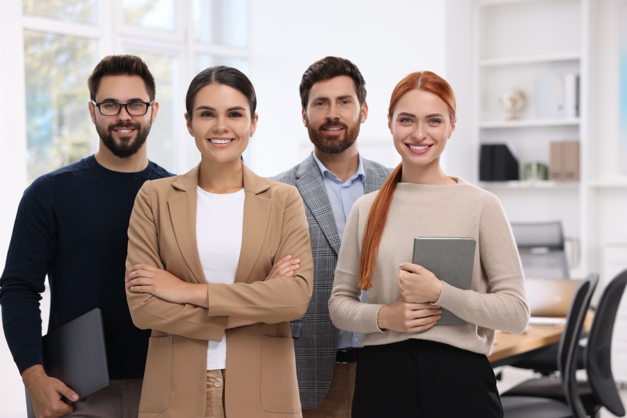 Four young professionals smiling and standing together in a bright office, two holding tablets and laptops.