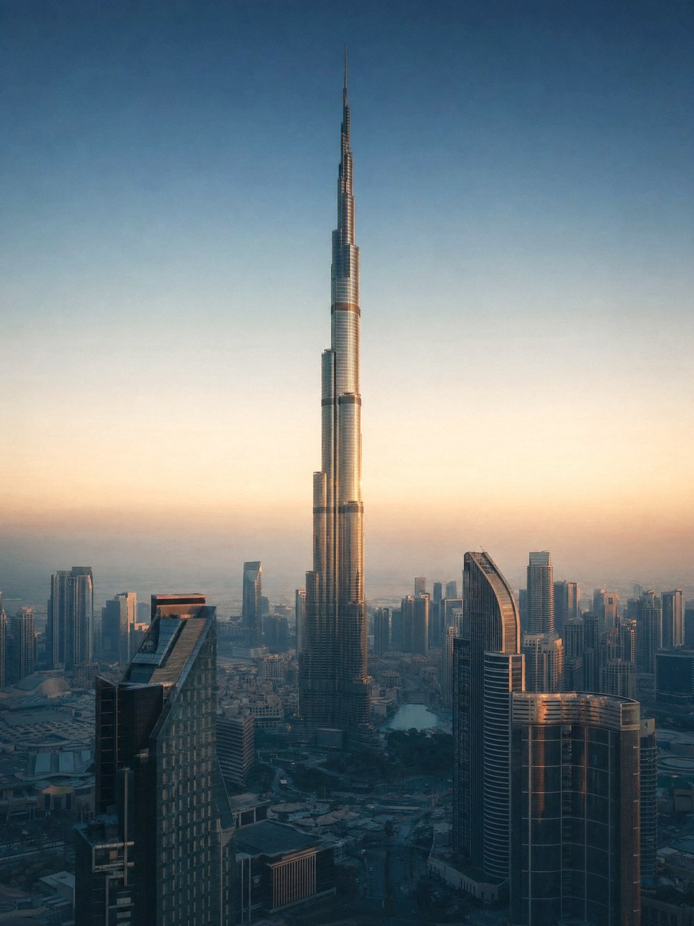 Rooftop view across Downtown Dubai at golden hour, the Burj Khalifa rising through desert haze