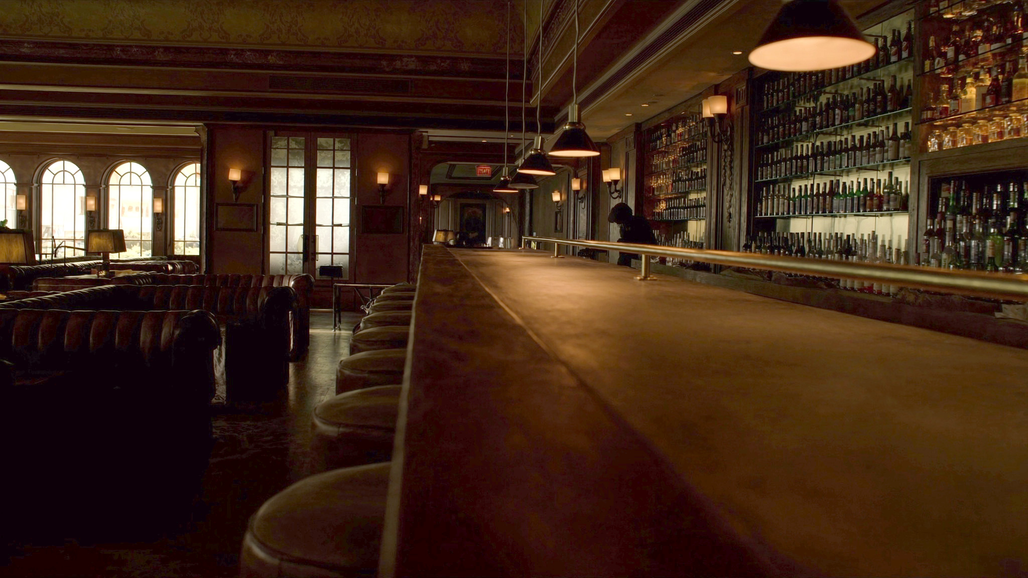 A quiet hotel cocktail bar in warm amber light, bottles on the back shelves and arched windows glowing at the far end