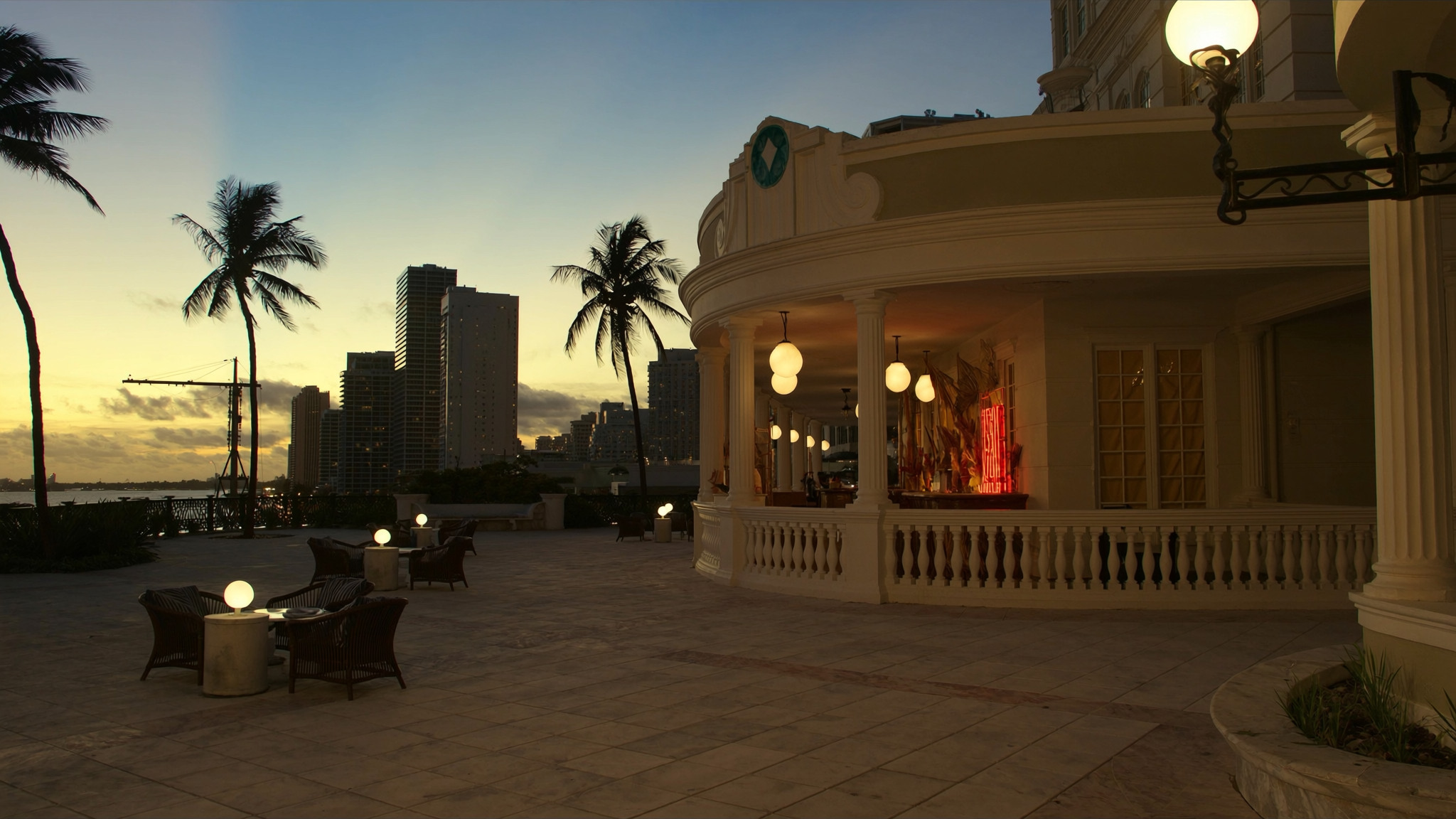 An empty hotel terrace in Miami Beach at dusk, globe lanterns along a colonnade, palms and the waterfront skyline beyond