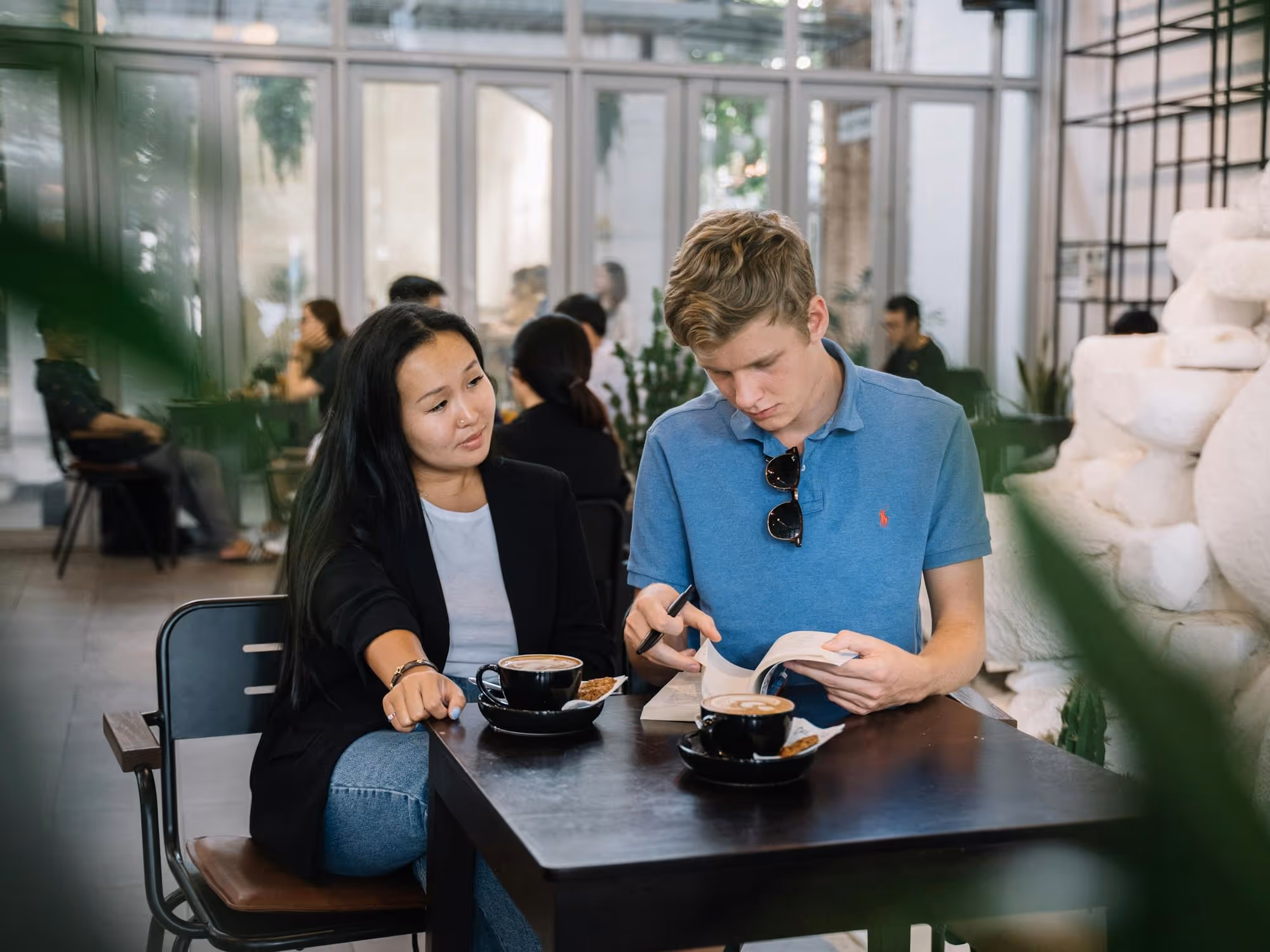 A young man reading a book and a woman looking at him while sitting at a café table with two cups of coffee.