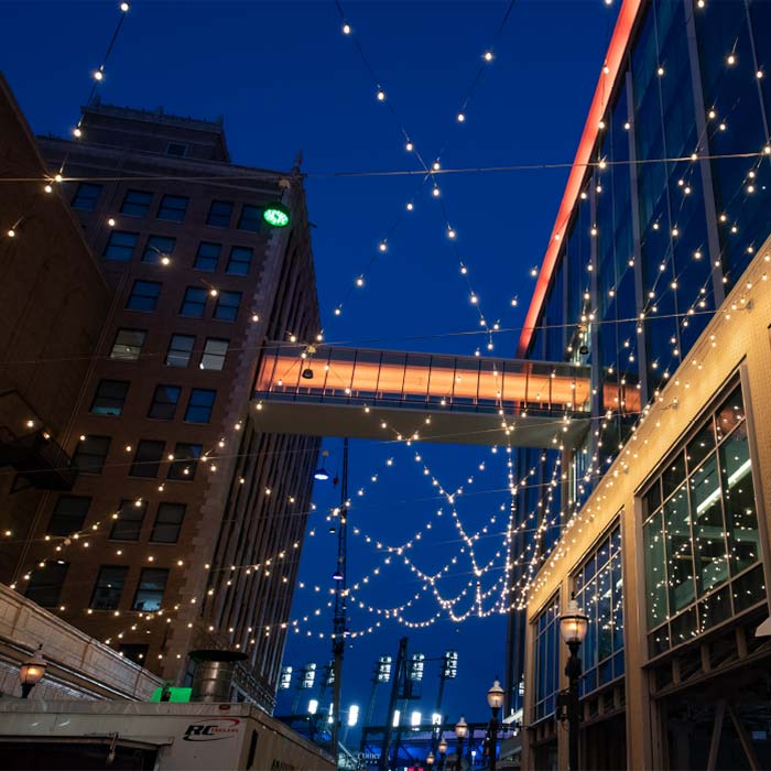Evening city street with string lights crisscrossing between buildings and illuminated skywalk.