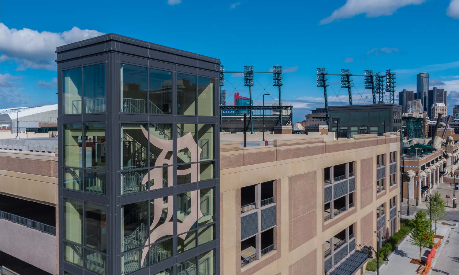 Exterior view of a modern building with a large glass elevator shaft displaying the Detroit Tigers logo, with stadium lights and city skyline in the background.
