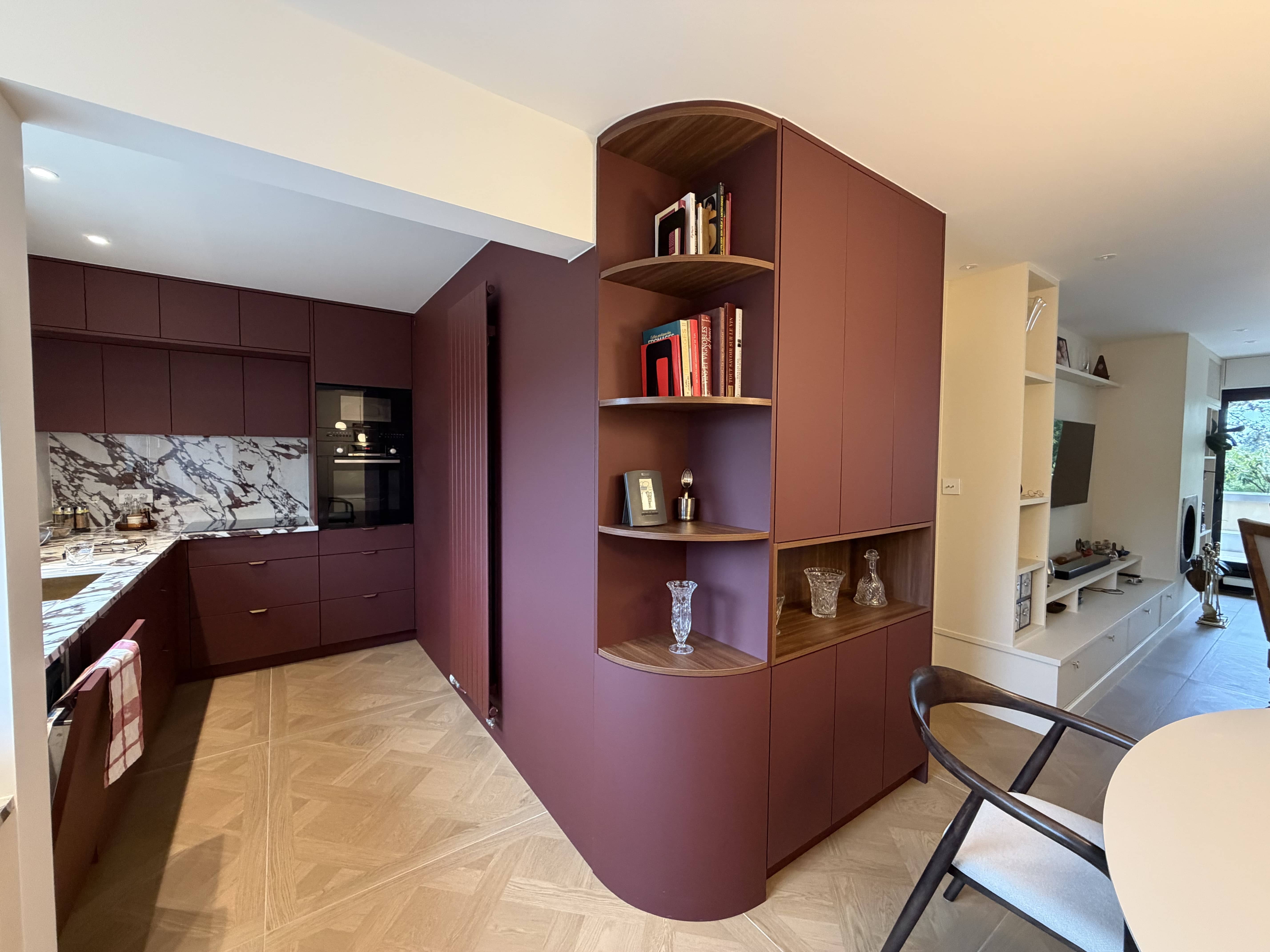 Modern kitchen and living area with burgundy cabinets, marble countertops, open shelves with glassware, and wood-patterned flooring.