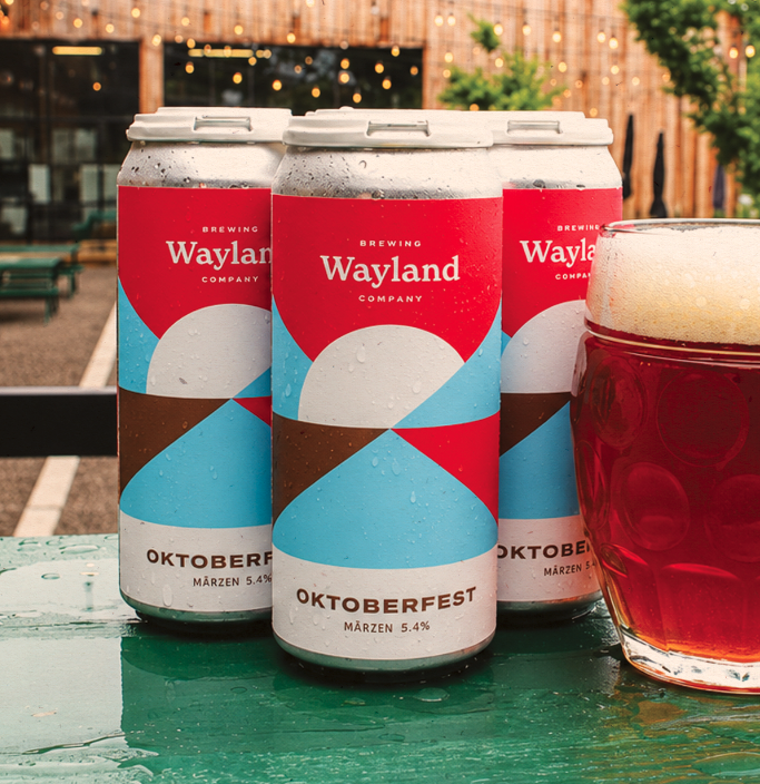 Three cans of Wayland Brewing Oktoberfest Märzen 5.4% beer with a glass of amber beer on a green table.