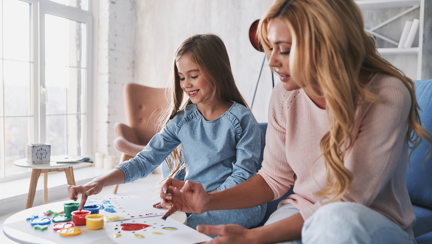 Smiling woman and young girl painting with watercolors together at a white table in a bright room.