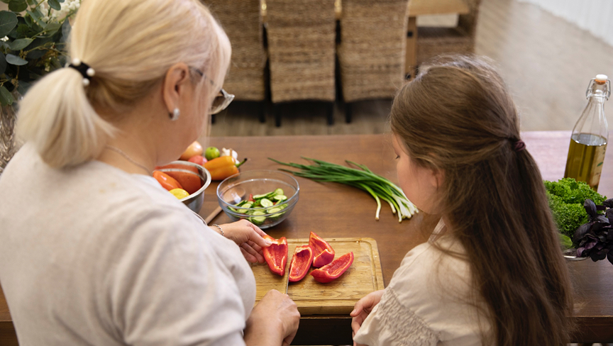 An older woman cutting red bell pepper slices on a wooden board while a young girl watches at a kitchen table with fresh vegetables nearby.