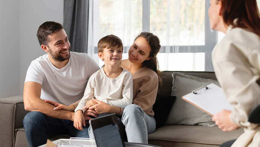 Parents sitting on a couch with their young son during a conversation with a professional holding a clipboard.