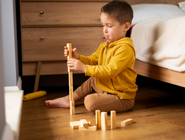 Young boy in a yellow hoodie sitting on the floor and stacking wooden blocks near a bed.