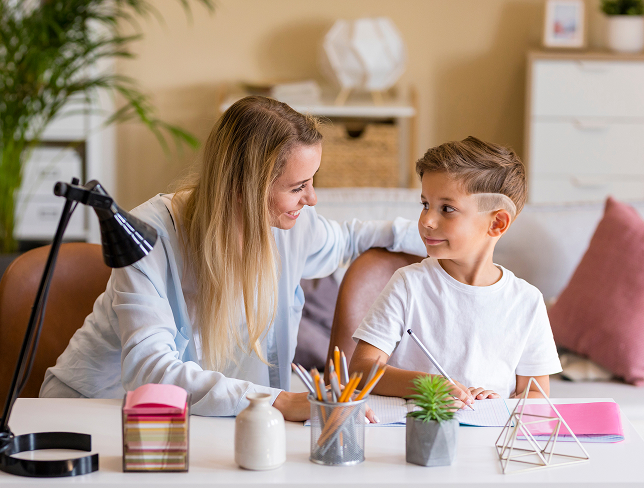 Woman smiling and engaging with a young boy as he writes at a desk in a cozy home setting.