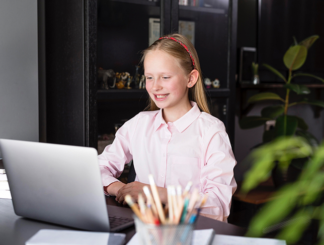 Smiling young girl wearing a pink shirt and headband sitting at a desk using a laptop in a room with a black cabinet and plants.