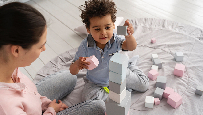 A child stacking pastel-colored wooden blocks while sitting on a round gray mat with an adult watching nearby.