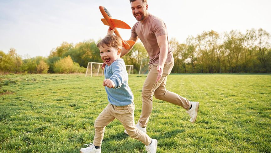 Smiling father and son running on green grass with the father holding an orange toy airplane.