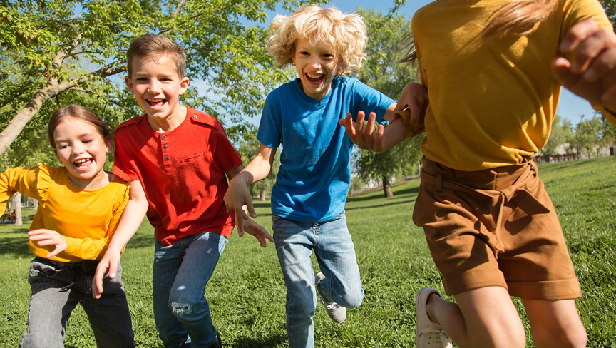 Four children holding hands and running on green grass in a sunny park.