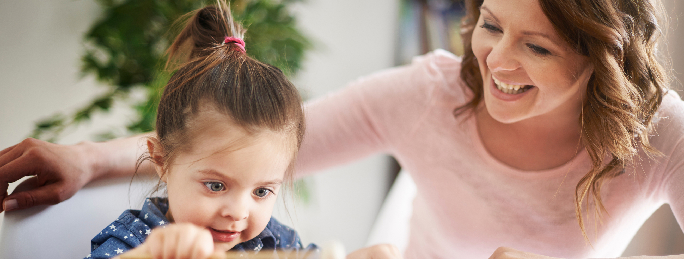 Smiling woman with curly hair watches a focused young girl with a ponytail playing or working on an activity.