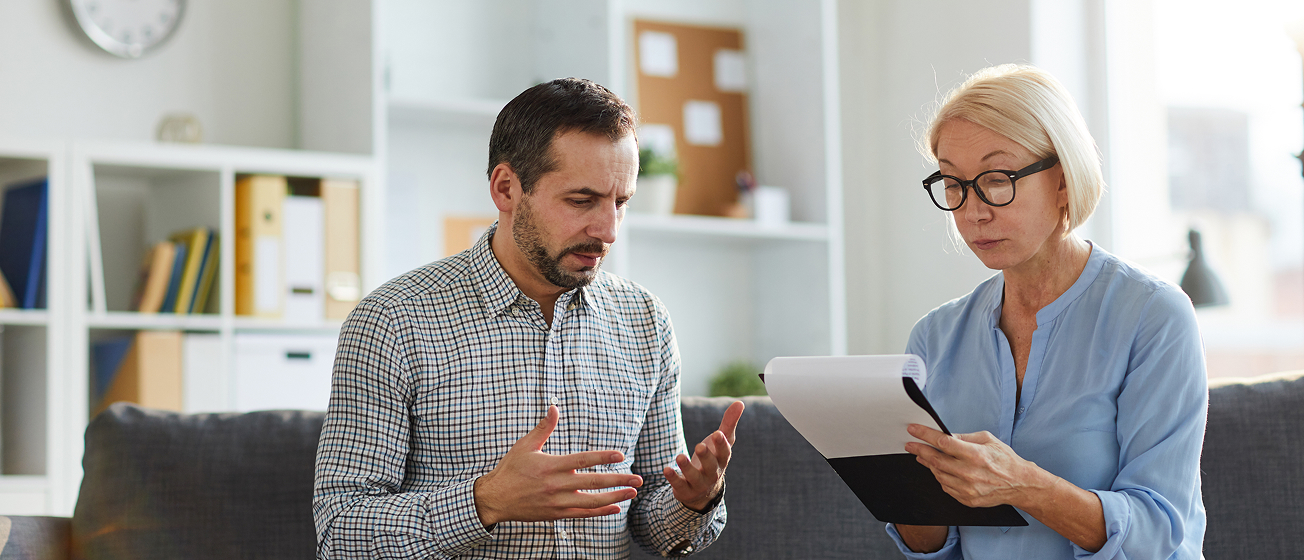 Man and woman seated on a couch having a serious discussion, the man gesturing with his hands and the woman looking at a clipboard with papers.