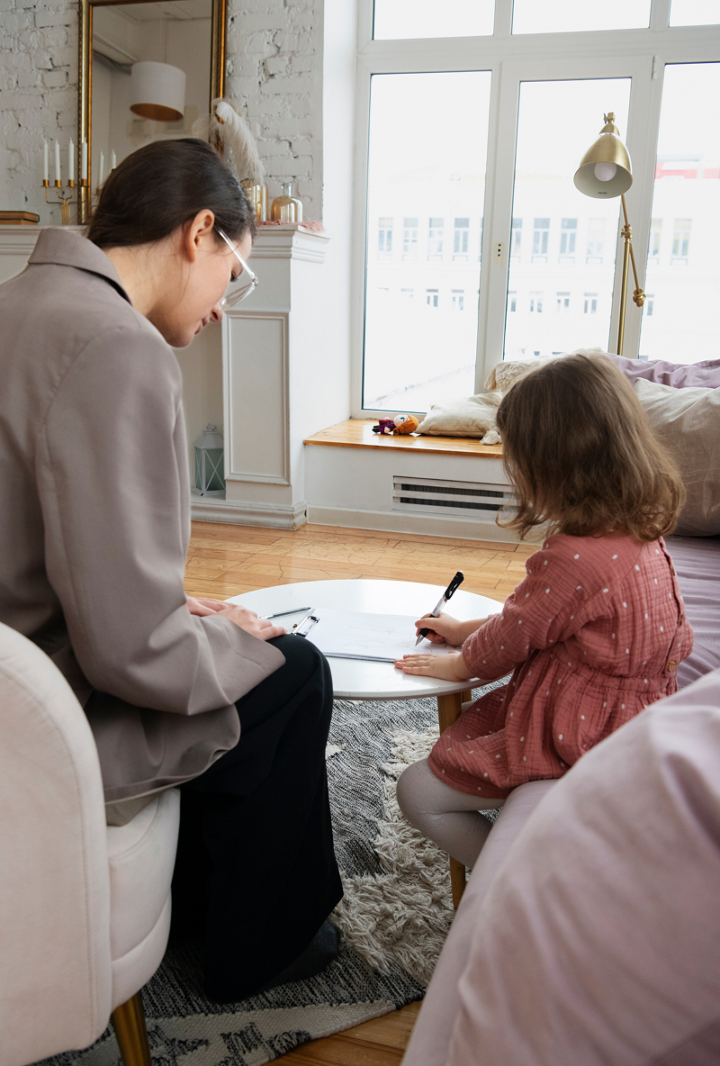 Woman and young girl sitting at a small round table in a bright room, the girl is drawing with a pen on paper while the woman watches.