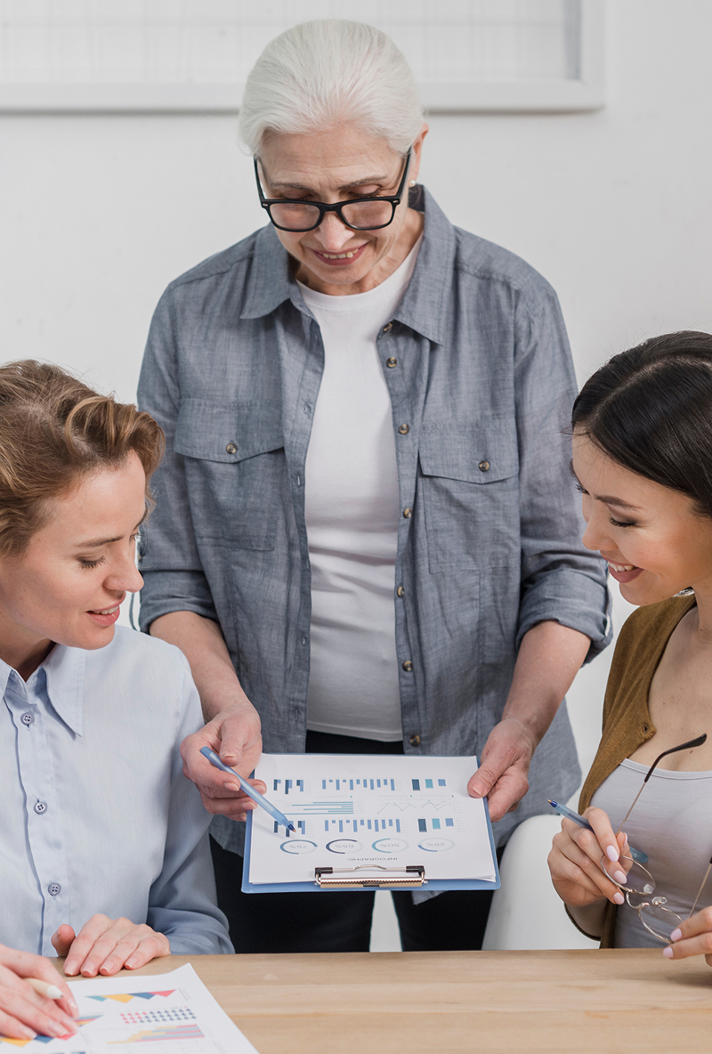 Three women reviewing charts and graphs on paper during a meeting, one standing and pointing at a clipboard.