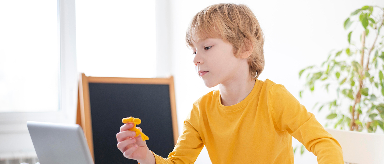 Young boy in a yellow shirt holding a yellow toy figure, sitting at a desk with a laptop in a bright room.