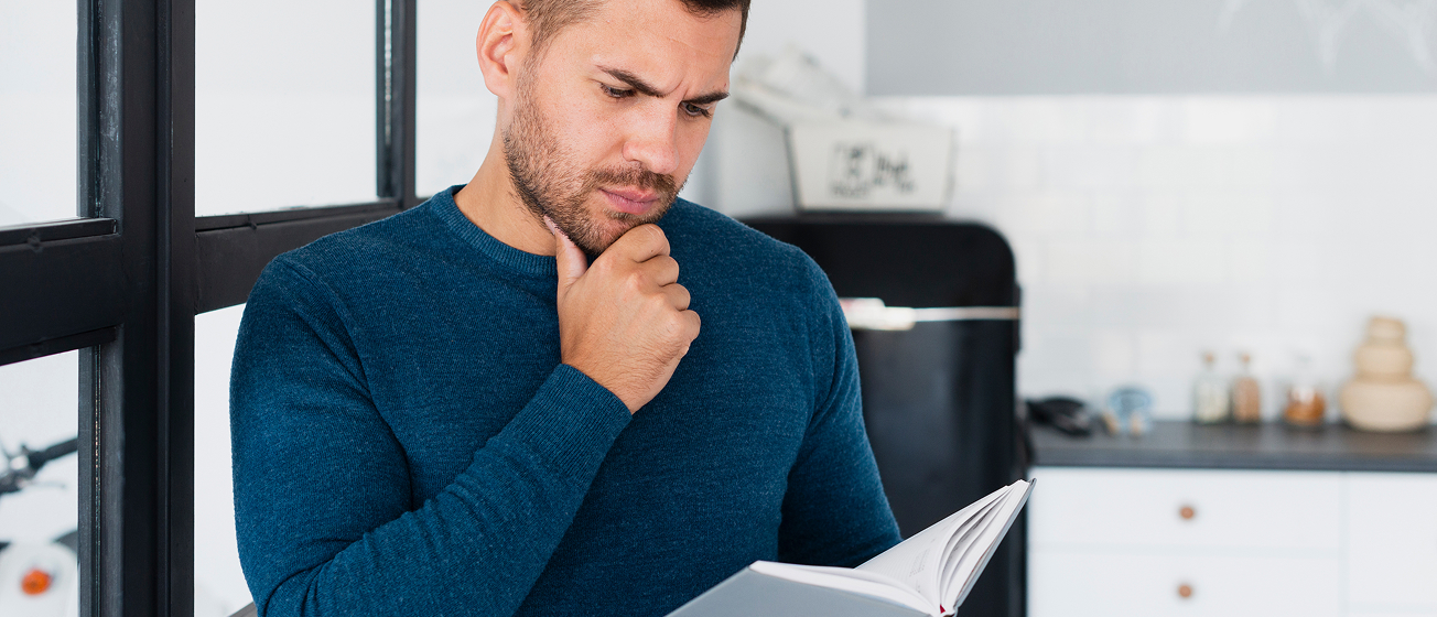 Man in blue sweater reading and thoughtfully looking at an open book in a modern kitchen.